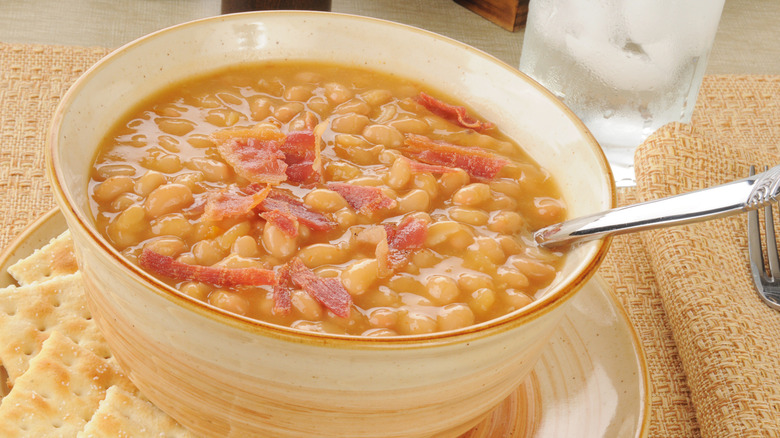 A bowl of bean with bacon soup in white bowl, soda crackers on the side.