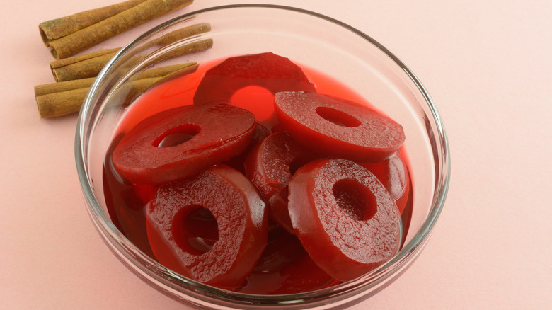 Red spiced apple rings in glass bowl with cinnamon sticks on pink background