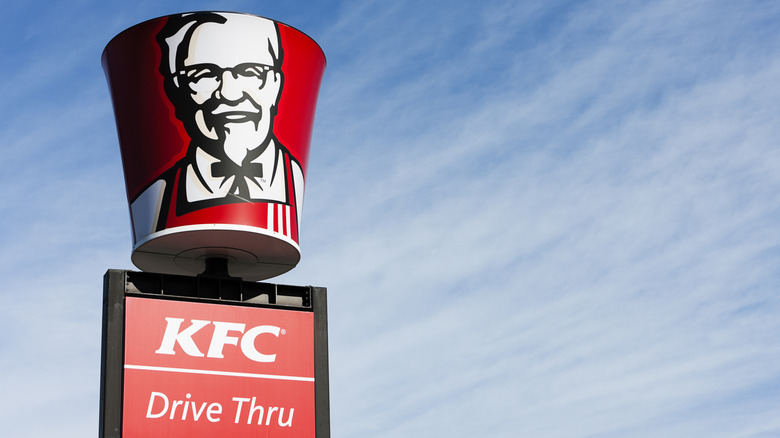 KFC sign, topped with rotating bucket with Colonel Sanders' face, against blue sky with wispy clouds.