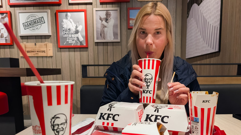 A blonde woman in a winter coat sips her soft drink from a red straw at a KFC in front of nostalgic black and white photos.