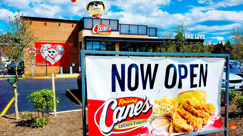 Photo of a new Raising Cane's location, partially hidden behind a large "now open" sign.