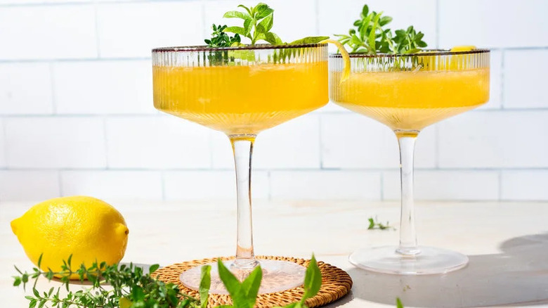 Two coupe glasses with yellow liqud and herbal garnishes next to lemon against white background.