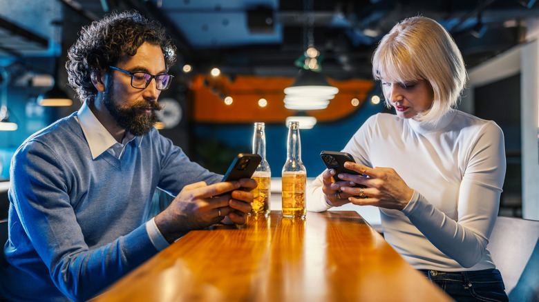 Disinterested, bored-looking people sitting on phone at bar with beers