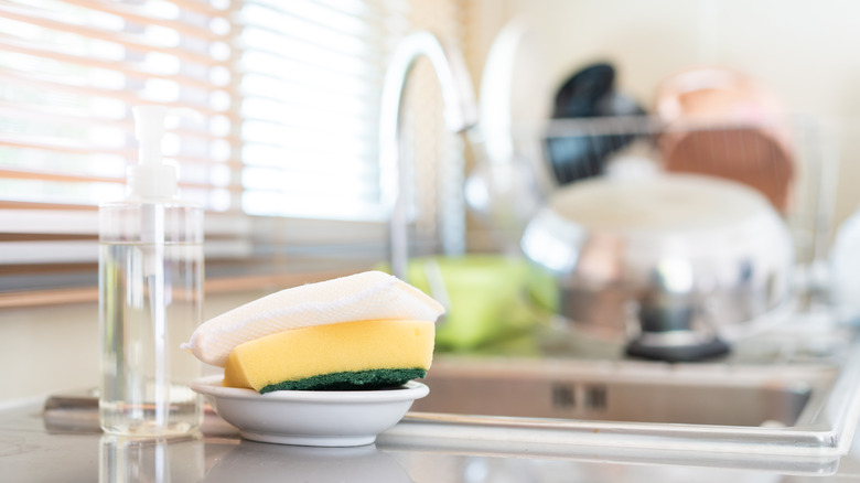 Scrubber sponges in a small white dish, in focus beside a transparent pump bottle of clear soap, with a sink, faucet, and stack of clean dishes out of focus in the background