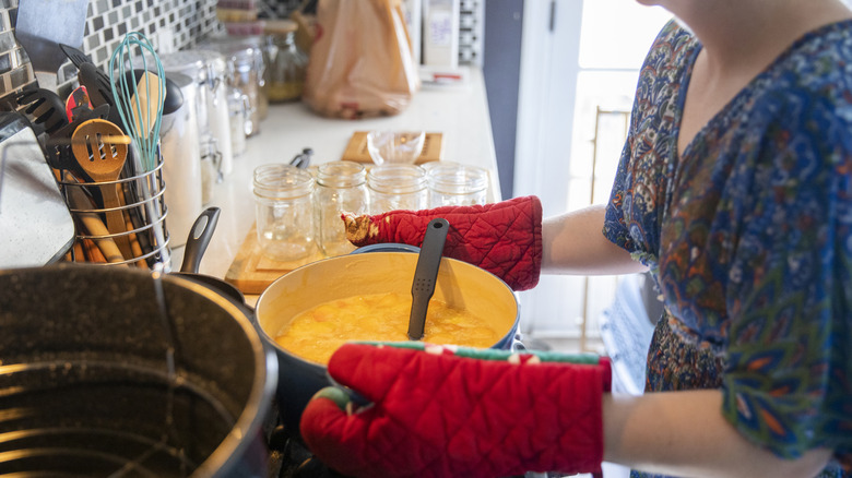 Woman in a blue, floral/paisley print dress and oven mitts, about to lift a Le Creuset Dutch oven full of homemade jam from the stovetop, with her canner visible at bottom left and prepared mason jars beside her on the counter