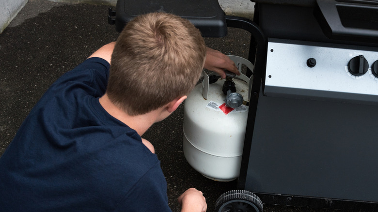 male teen in a blue T-shirt turning the valve of a propane tank attached to a black grill