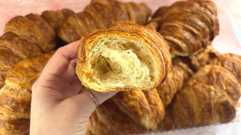 Hand holding bitten Costco croissant over box of croissants on pink surface