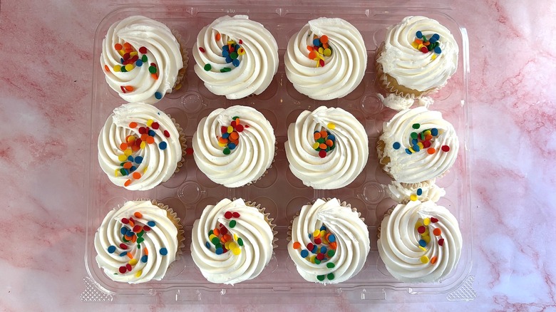 Overhead shot of Costco container of white cupcakes with white frosting on pink surface