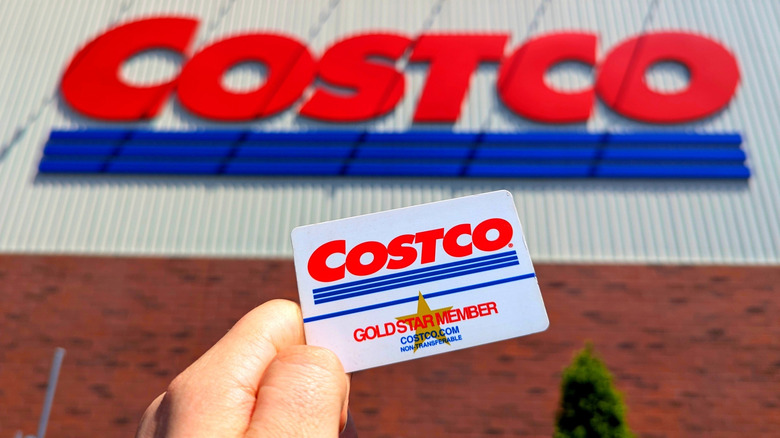 Hand holding Gold Star Costco membership card outside in front of Costco building sign.