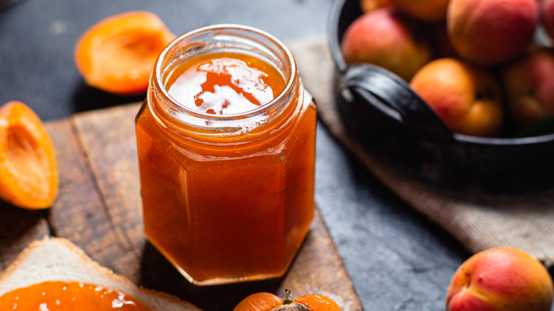 Ripe apricot jam in a glass jar on a table next to whole and halved apricots