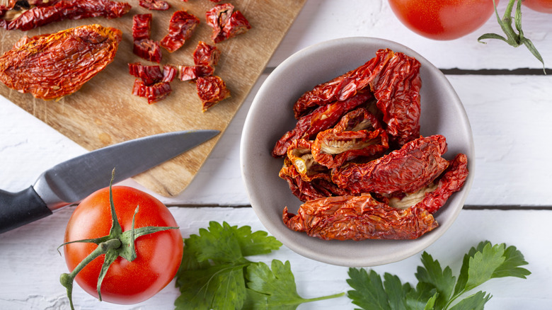 Bowl of sun-dried tomatoes with some being chopped on wooden cutting board
