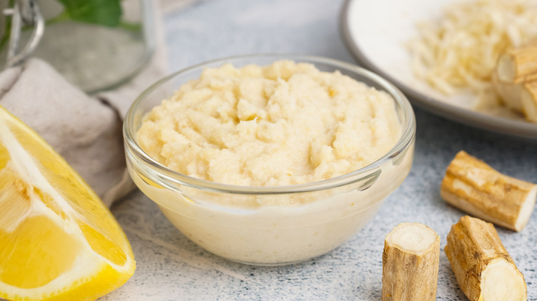 Prepared horseradish in a bowl next to horseradish root and lemon wedge