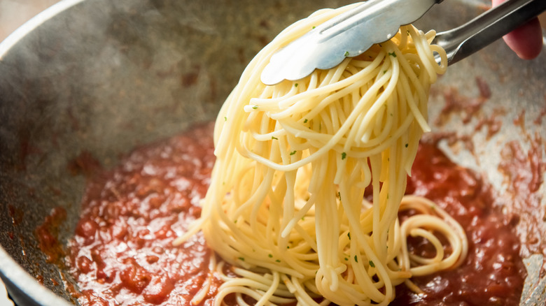 Tongs adding cooked spaghetti to pan of tomato pasta sauce