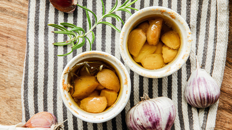 Garlic confit in two white bowls next to fresh garlic bulbs and sprig of rosemary