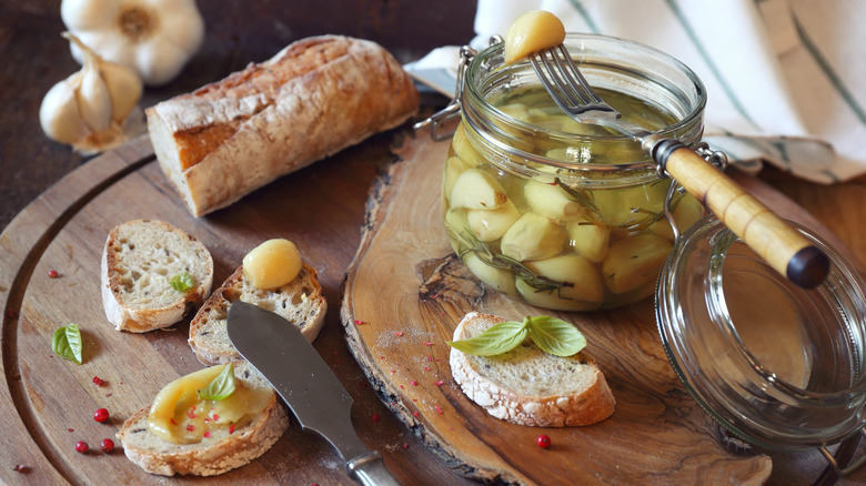 Jar of garlic confit, with cloves being spread onto slices of crusty baguette
