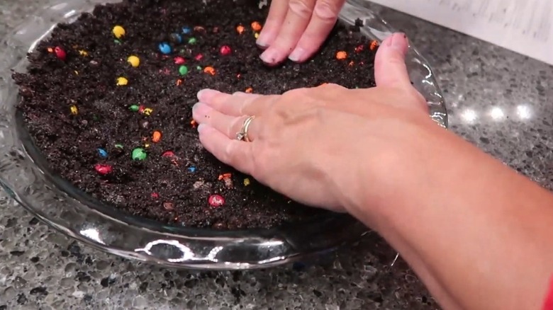 Hands pressing Oreo and M&M pie crust into glass pie dish