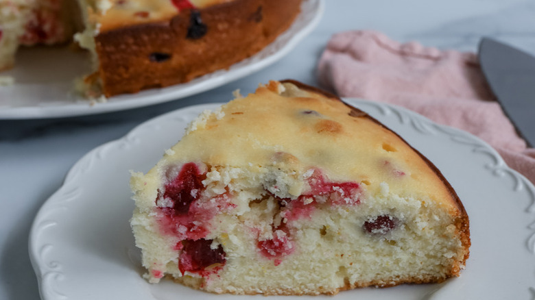 Slice of cranberry ricotta cake on white plate with the whole cake in the back