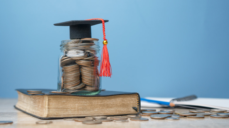 Small jar full of coins topped with tiny graduation hat and red tassel, all sitting on top of a book, with more coins scattered around the ground