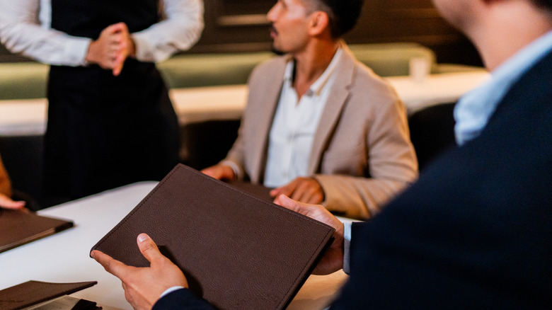 A close-up of man holding a menu at a restaurant with another diner and a server in the background