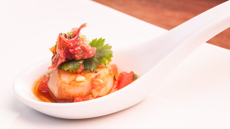 A close-up of a seared scallop served in a spoon with herbs and a garnish