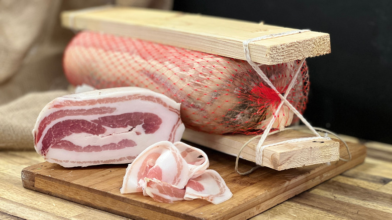 whole pancetta and a thin slice on a wooden cutting board