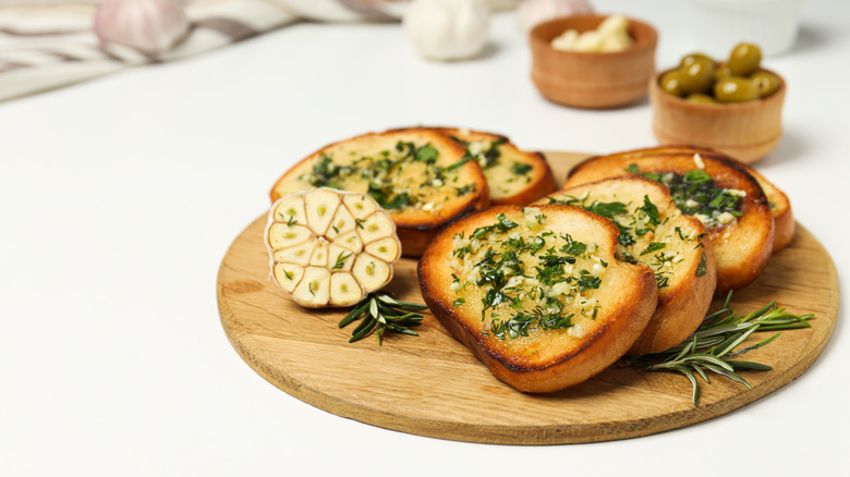Mini toasts spread with garlic herb butter on cutting board with rosemary and garlic bulb cut in half