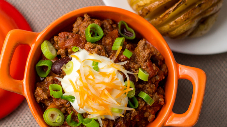 Bowl of chili wth sour cream, shredded cheese, and chives with a sliced baked potato in the background