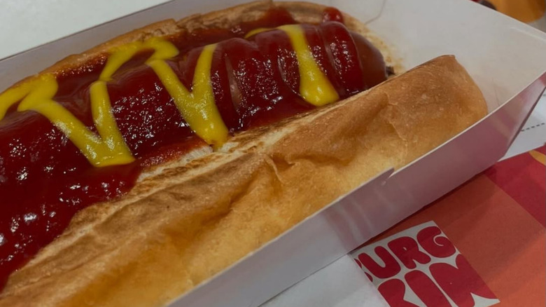 Close-up image of a Burger King grilled hot dog, with ketchup and mustard, in an open-topped cardboard container.