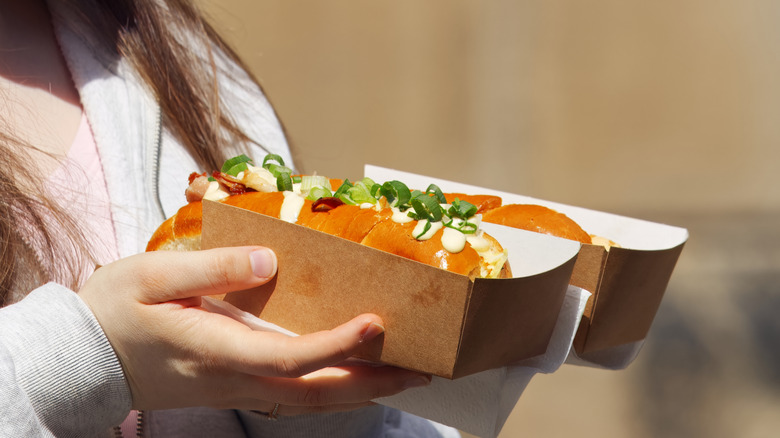 Person holding two loaded hot dogs in brown cardboard takeout packaging.