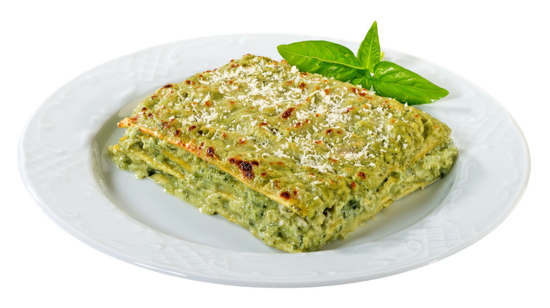 A slice of spinach lasagna on a white plate with basil on a white background