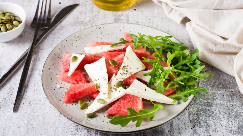Watermelon, feta & arugula salad on a plate with utensils, oil, and a kitchen towel