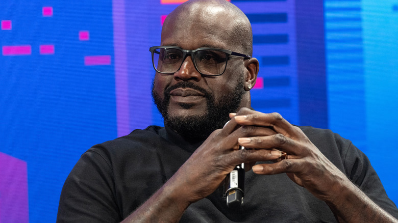 Shaquille O'Neal against blue background with hands clasped in front of him holding a microphone