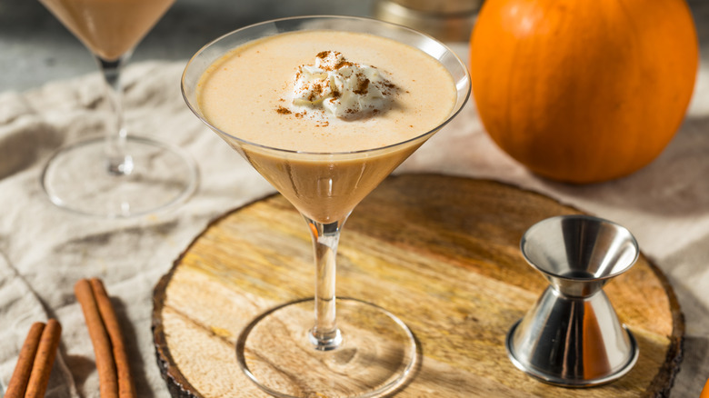 A pumpkin martini with a dollop of whipped cream on a table with pumpkins and cinnamon sticks