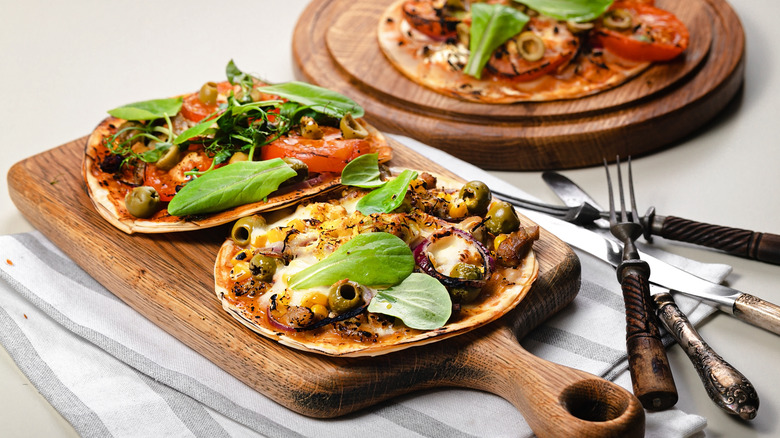 Three tortillas on wooden serving trays topped with olives, tomatoes, and leafy greens