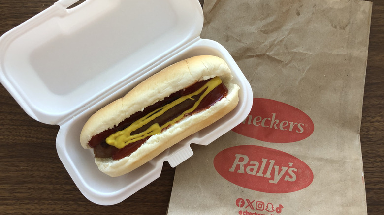 Rally's grilled hot dog with mustard in styrofoam box with to-go bag.