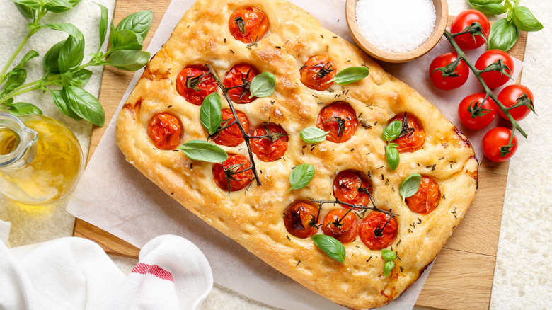 Focaccia with tomatoes, spices, and oil on wooden board with fresh tomatoes, salt, and oil