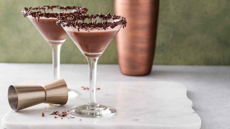 two Chocolate martini with chocolate sprinkles on rim of glass, against green wall with cocktail shaker behind