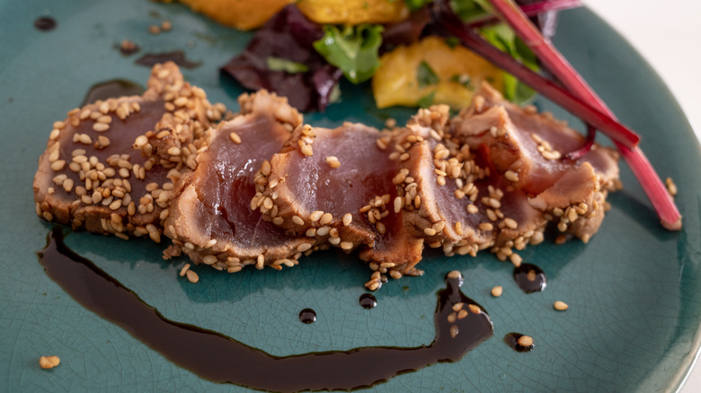 small slices of seared tuna, crusted with sesame seeds, served on a deep blue-green plate with light vegetables and a decorative drizzle of what appears to be soy sauce