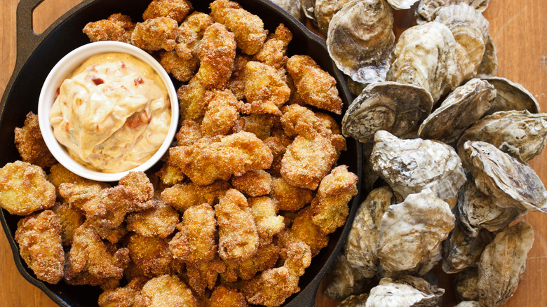 a mound of fried oysters served in a cast-iron skillet, with a ramekin full of dipping sauce, and with live, in-shell oysters mounded alongside