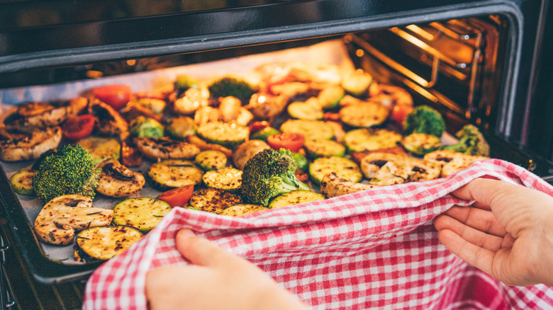 woman's hands sliding a tray of seasoned summery vegetables (broccoli, mushrooms, zucchini, cherry tomatoes) into a small oven