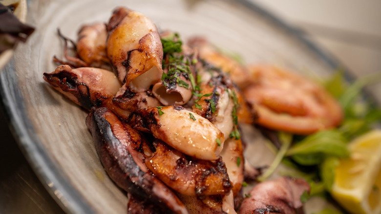 grilled baby squid on a rustic plate, with vegetable garnishes and a lemon wedge visible but out of focus behind them