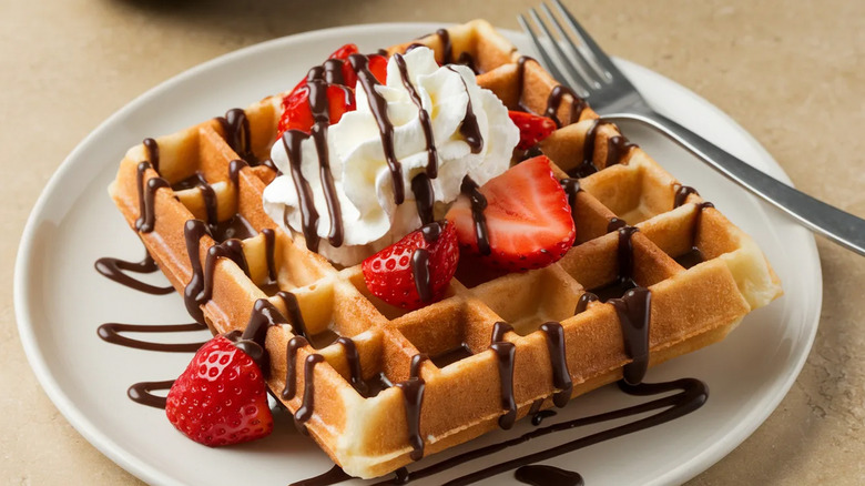 Belgian waffle topped with whipped cream, strawberries, chocolate drizzle on white plate with fork.