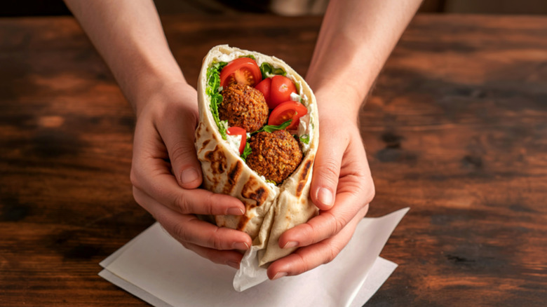 A person holds a falafel wrap over a napkin on a wooden table.