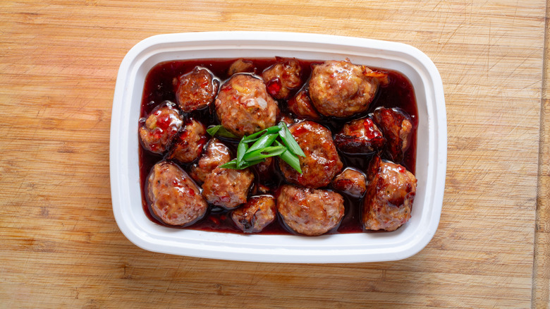 A white plastic container of grape jelly meatballs on wooden table.