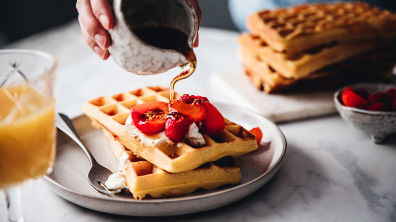 Close-up of a hand pouring maple syrup onto waffle topped with fruit.