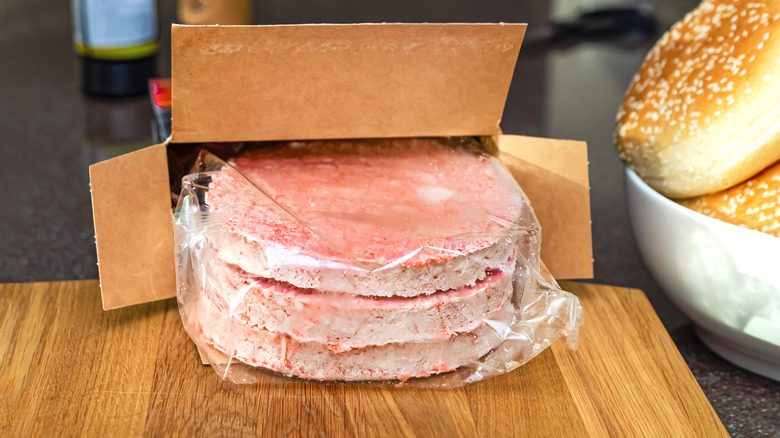 opened box of frozen burger patties on a cutting board, with three still-wrapped burger patties visible at the open end, with sesame-seed buns in a bowl alongside the cutting board