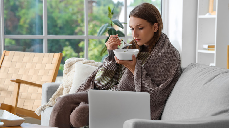 Woman wrapped in blanket eating soup on couch