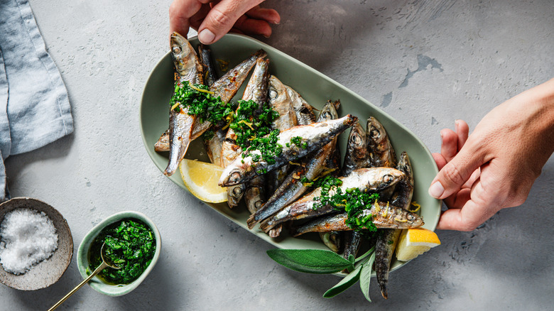 hands placing down an oval platter with grilled sardines, herbs and lemons