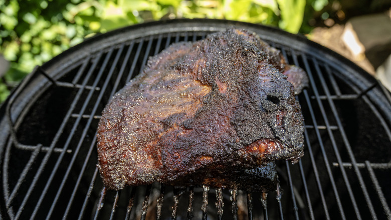 A well seasoned brisket cooking on a black kettle grill with blurred green grass in the background