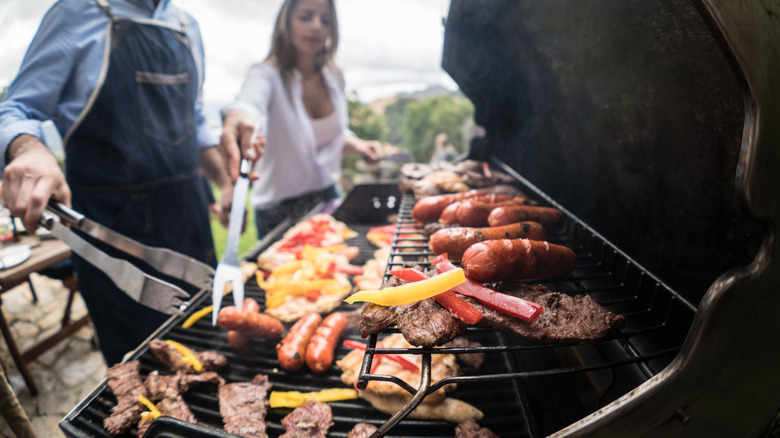 A couple of people are standing over a large grill covered in sausages, meat, and veggies with an outside environment blurred in the background